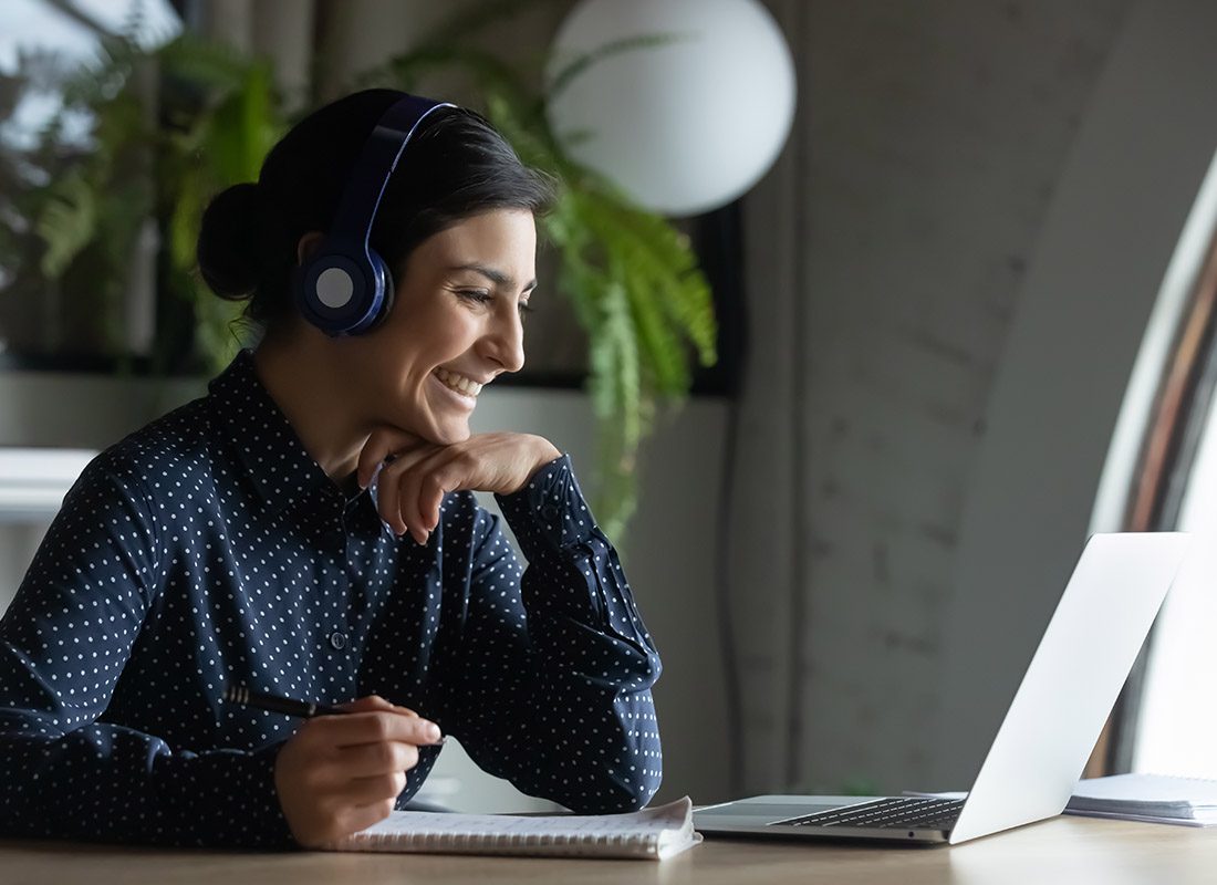 Join Our Webinar - Woman Smiling with Her Headset on as She Takes Notes While Watching a Webinar in Her Office at Her Desk